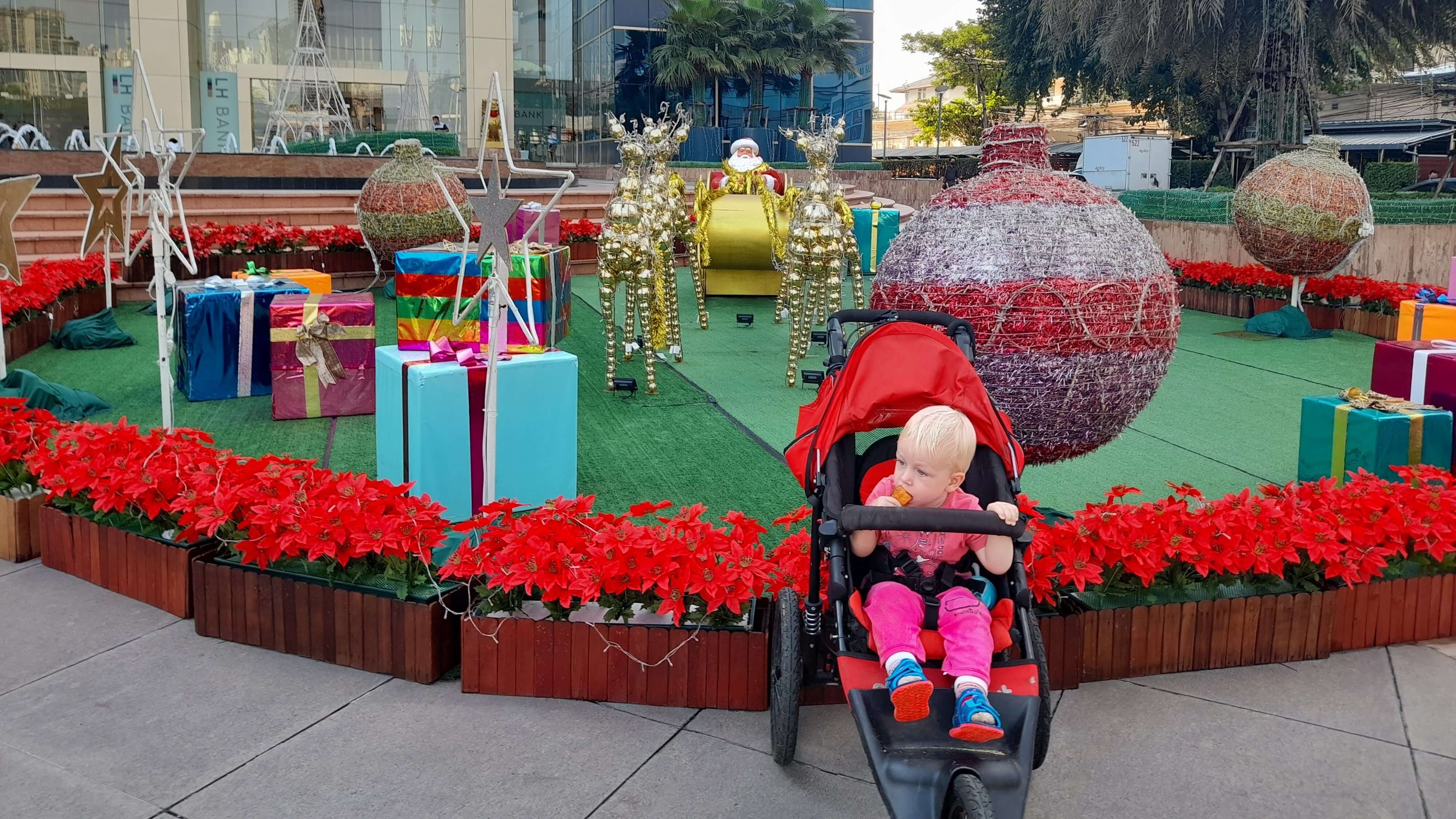 A toddler seated in a stroller in front of large Christmas decorations outside a Bangkok mall.