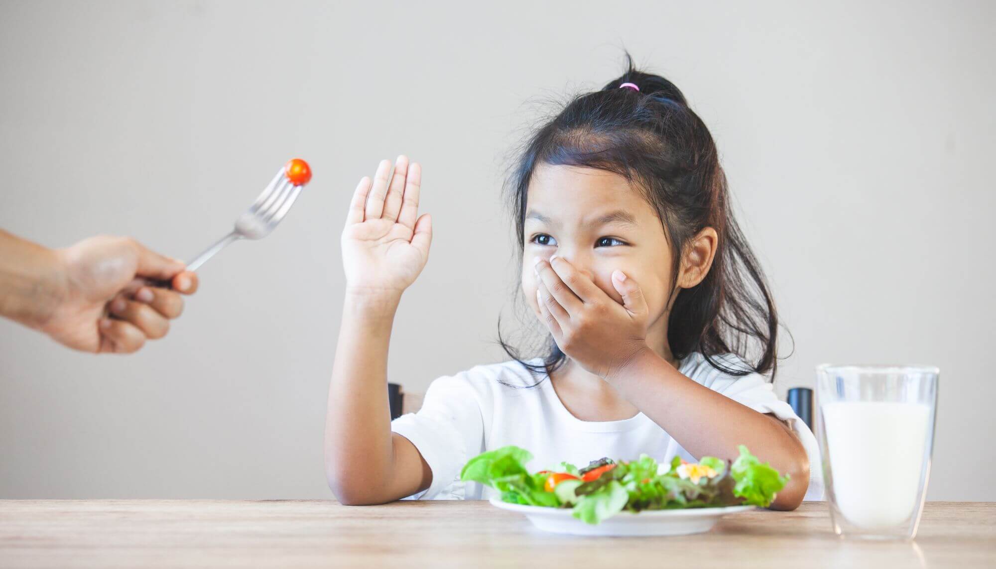 A four-year old Asian girl refusing the cherry tomato being offered to her on a fork by an off-camera adult. She is holding one hand over her mouth and the other in front of the tomato to say "No".