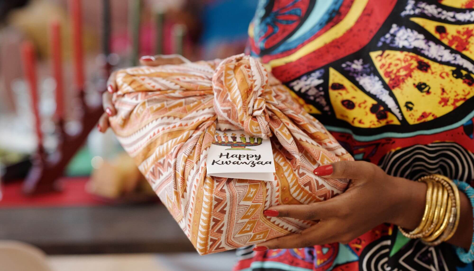 Hands holding a gift wrapped in colorful cloth with a label that reads "Happy Kwanzaa".
