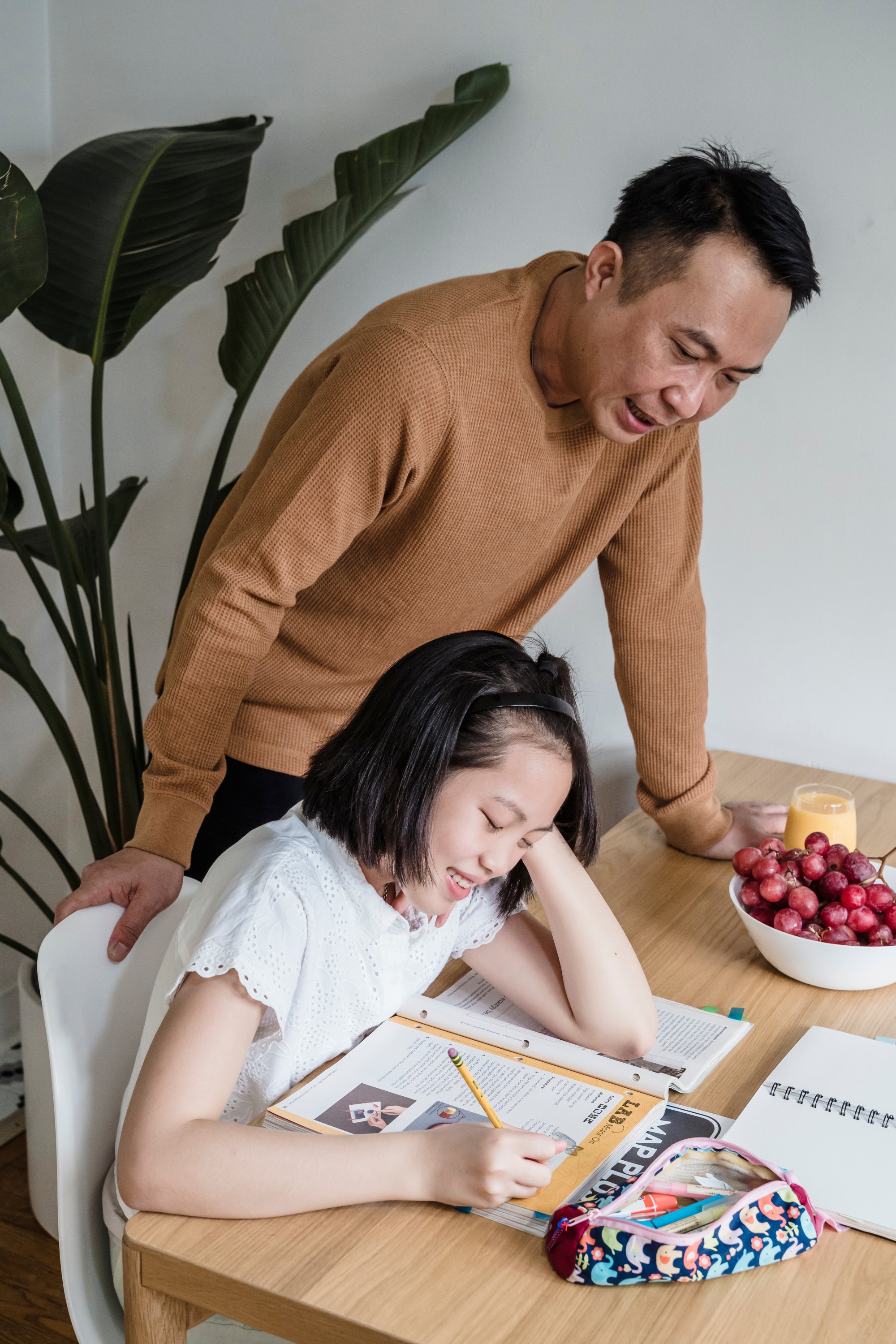 A child studies at a table with her father standing over her.