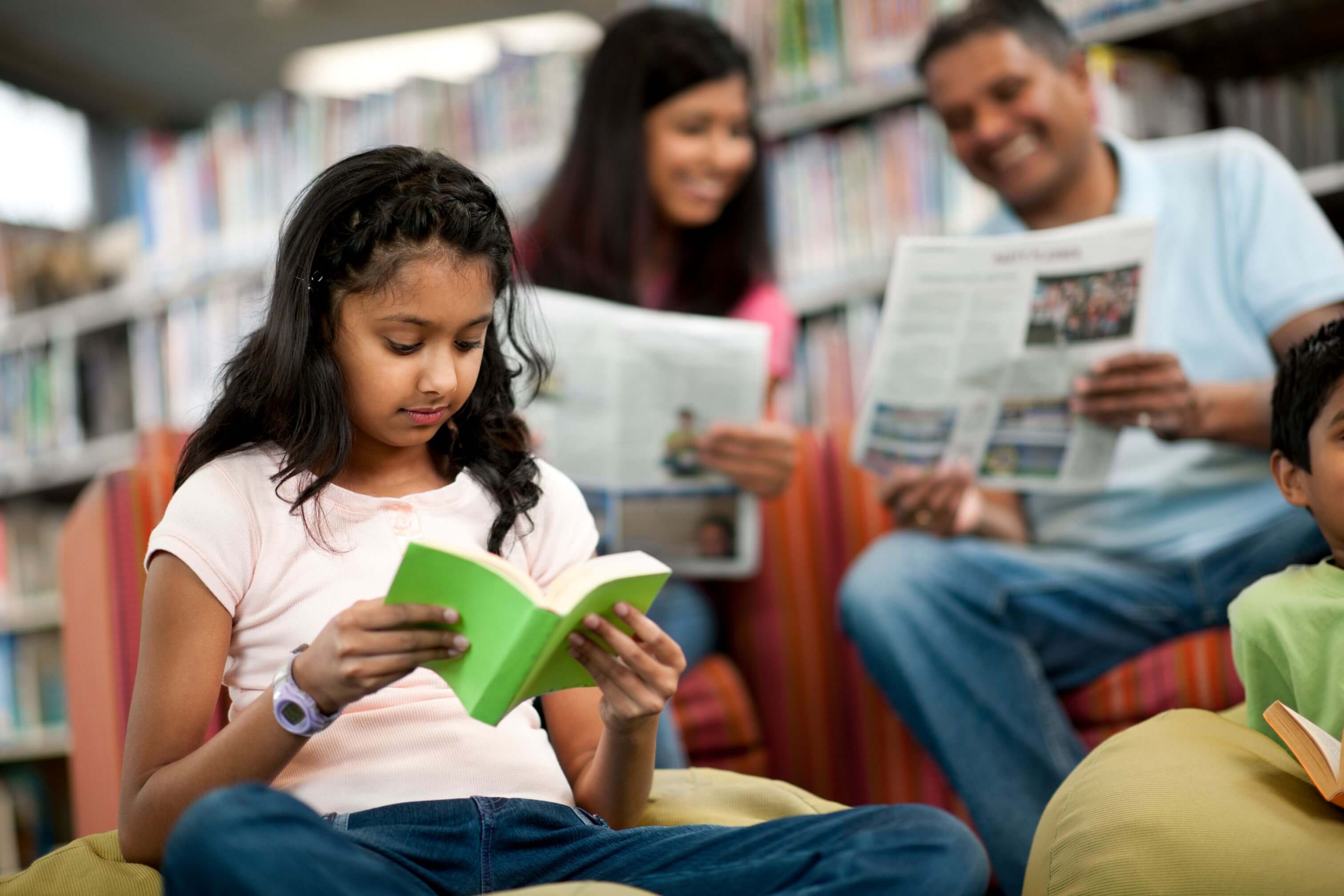 A girl reading a small book in the foreground and her parents reading newspapers in the background.