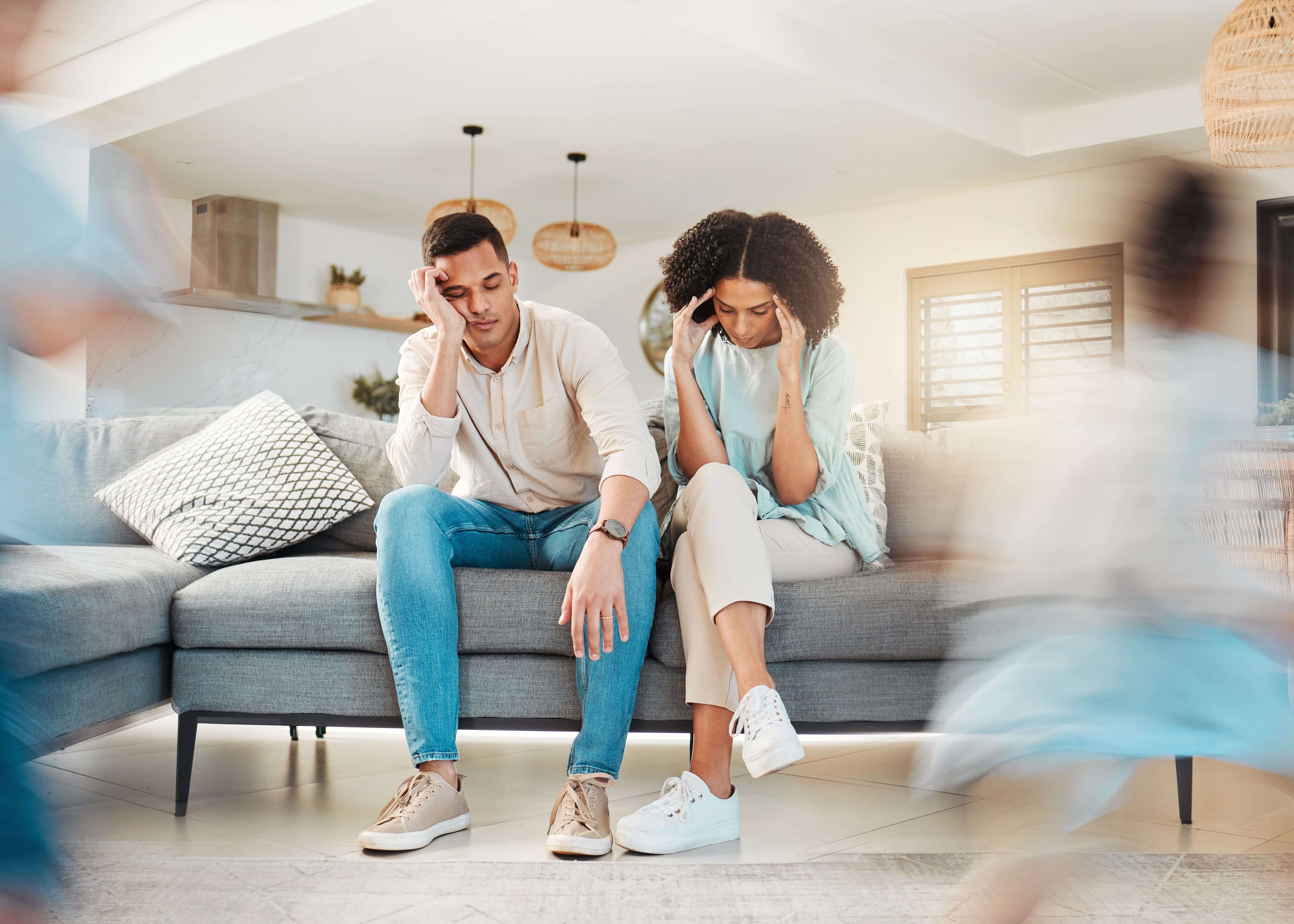 A Black mother and Black father sit on a sofa holding their heads exhausted as their children buzz around them