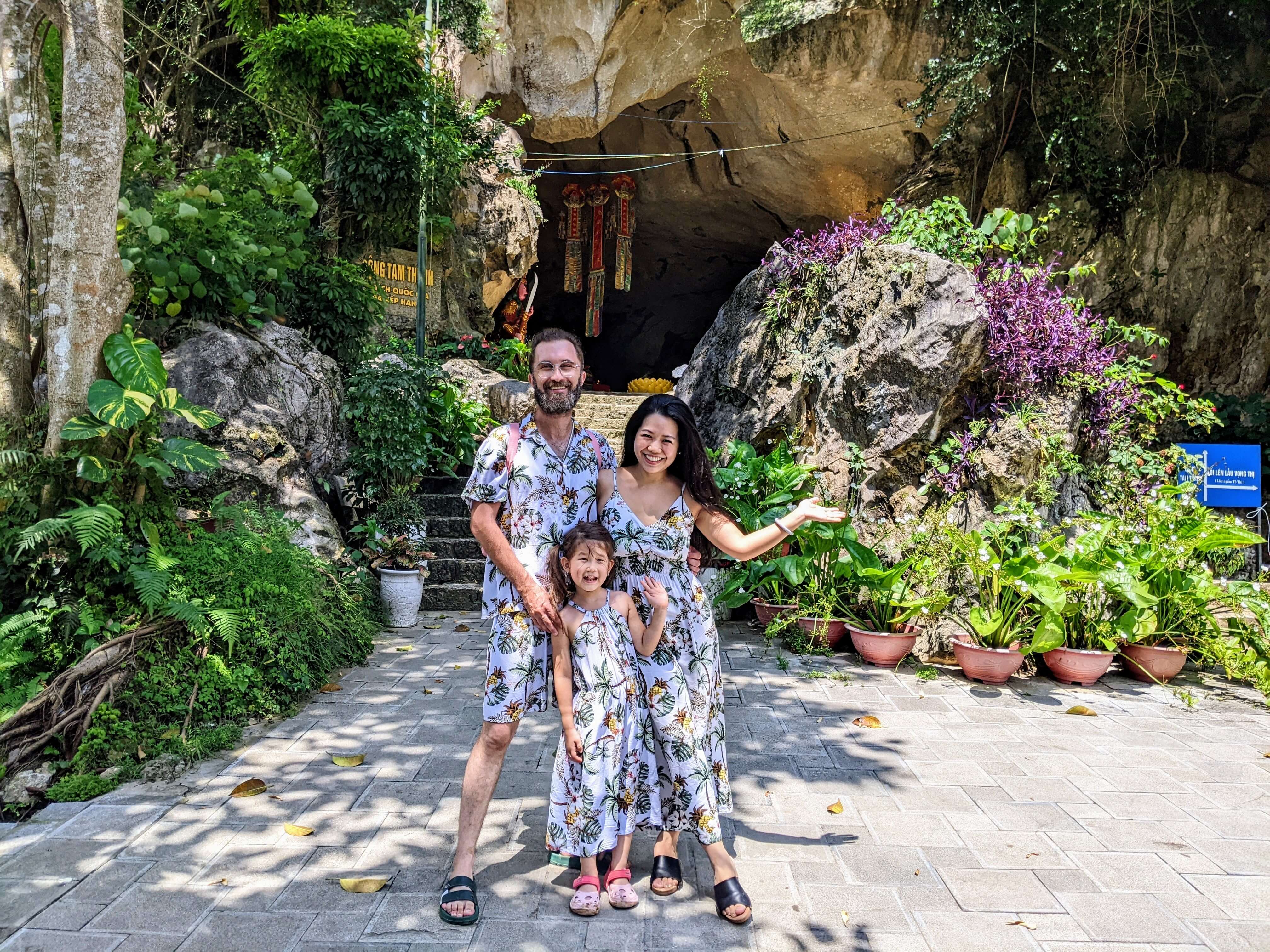 A family of three - a man, a woman, and their daughter - standing together in a garden, dressed in matching clothes and smiling at the camera.