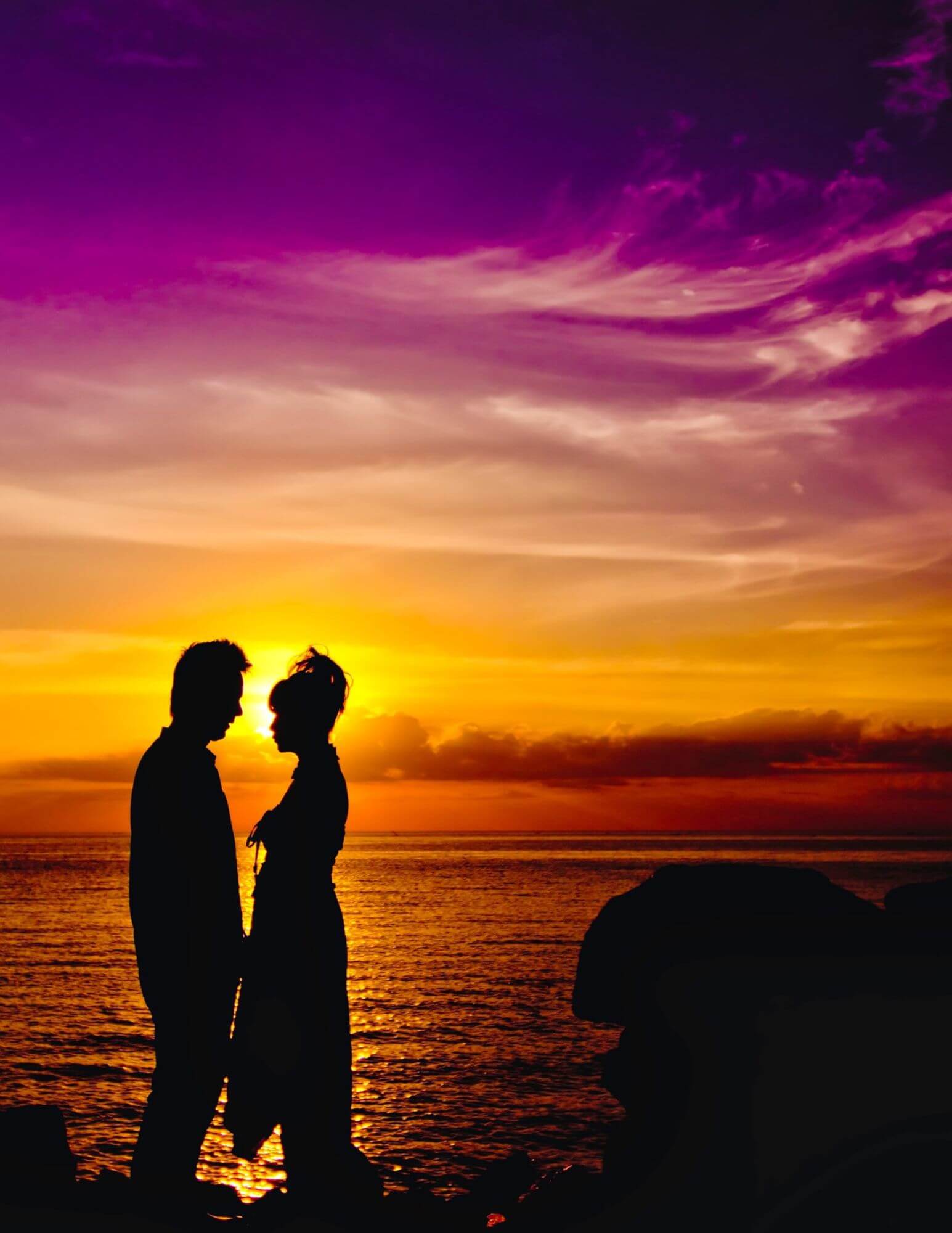 The silhouettes of a man and women standing close together and facing each other in front of a purple and orange sunset