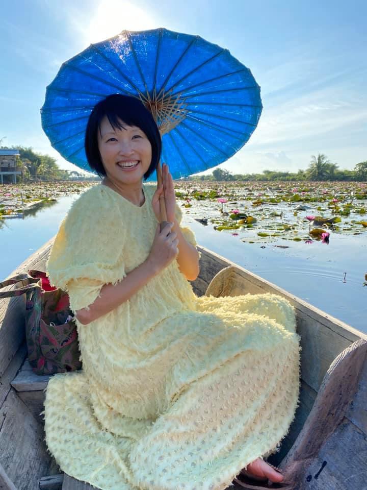 Cecilia, a Chinese woman in her forties, sitting in a wooden boat on a lotus pond holding a blue parasol and smiling