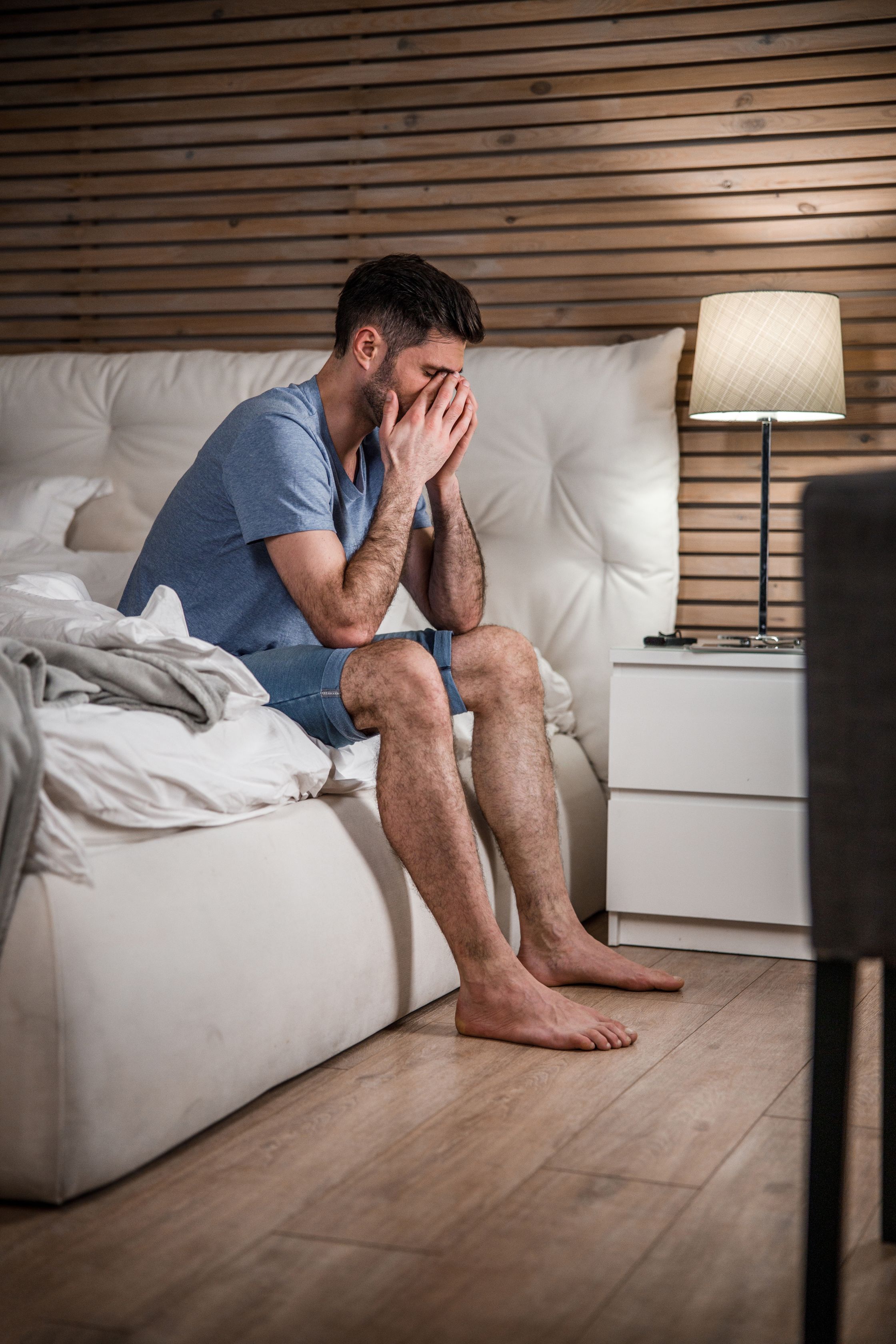 A white man sitting on the edge of a bed and rubbing his tired eyes