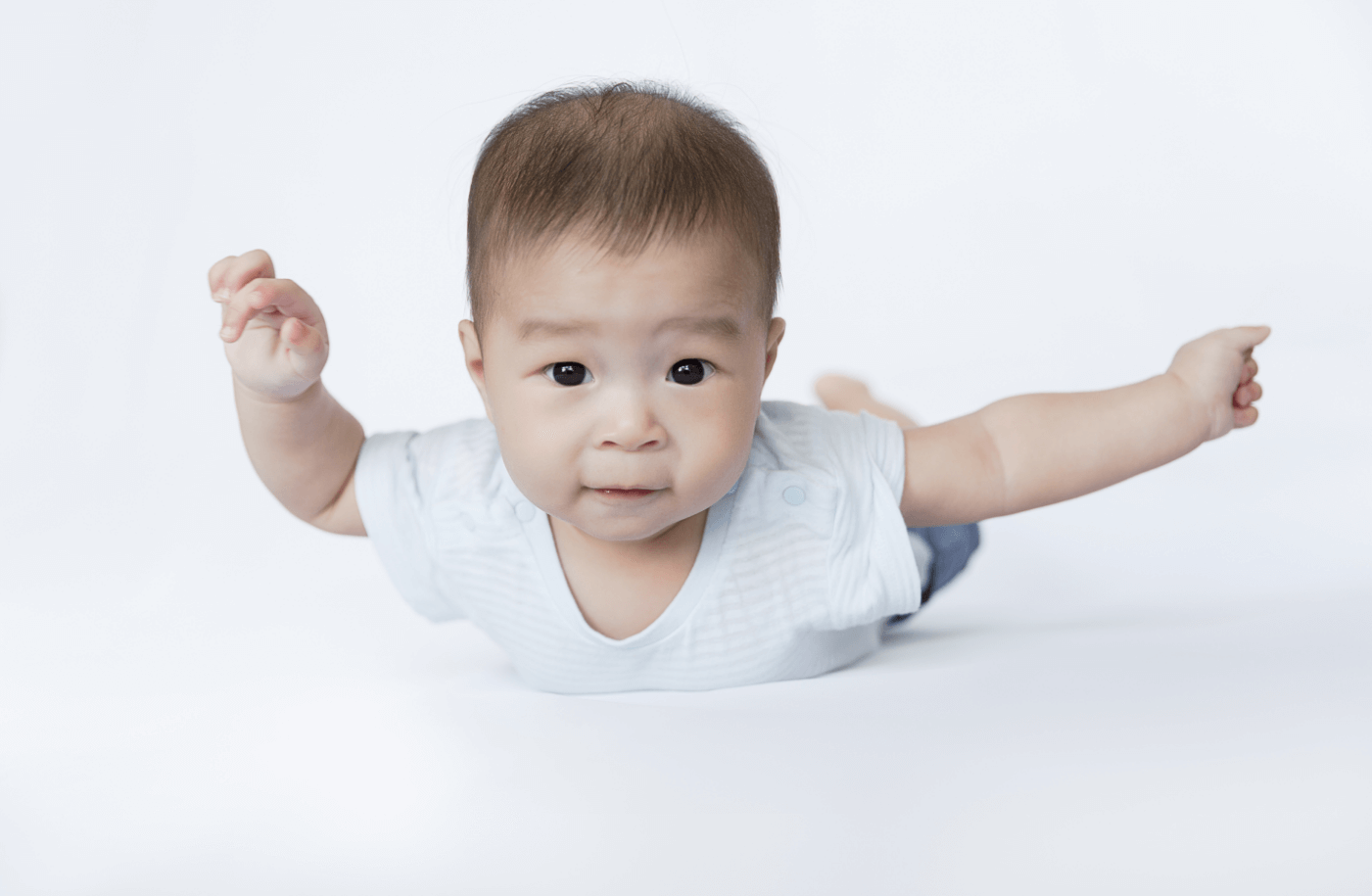 An Asian baby balancing on their tummy, arms spread out like an airplane, looking directly at the camera.