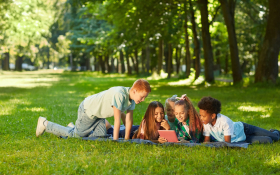 A racially diverse group of teen and tween girls and boys lying on a picnic blanket looking at an iPad. 
