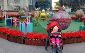 A toddler seated in a stroller in front of a large display of Christmas decorations outside a Bangkok mall.