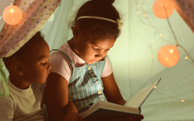 A girl and boy sitting inside a play tent filled with fairy lights and reading together