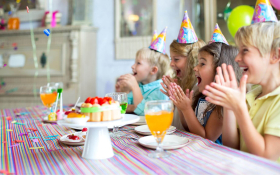 A group of children sitting at a a party table. They are wearing party hats and smiling and clapping.