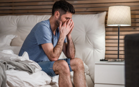 A white man sitting on the edge of a bed and rubbing his tired eyes