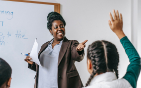 A teacher stands at the front of a class whilst a student raises her hand.