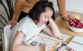 A child studies at a table with her father looking over her.
